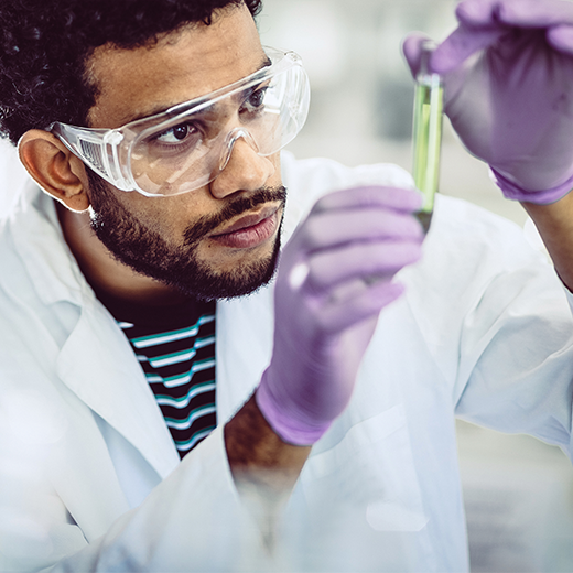 A man wearing a lab coat, gloves, and safety glasses works with a vial of liquid.